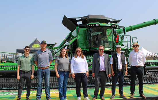 Group of seven attendees standing in front of a large green combine harvester at an outdoor equipment event, posing for a photo in front of the machine.