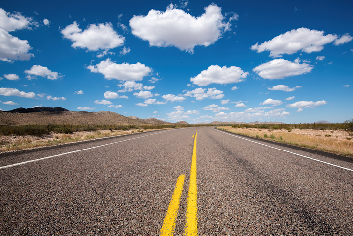 Two-lane highway stretching through a desert landscape beneath a bright blue sky filled with scattered white clouds.