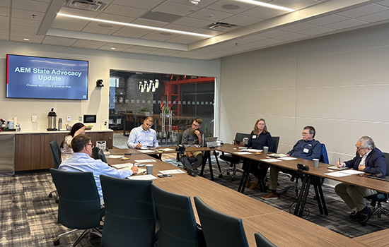 Small group meeting in a modern conference room with participants seated around a table, reviewing documents during an “AEM State Advocacy Update” displayed on a wall-mounted screen.