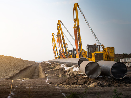 Large pipeline construction site with multiple yellow sideboom cranes positioned along a trench, lifting and aligning sections of steel pipe in a rural landscape.